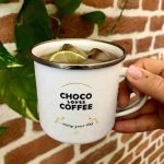 Bílý hrnček s nápisem Choco Loves Coffee a ledovým nápojem uvnitř, držený v ruce.