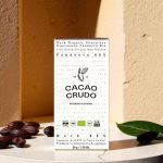 Balení tmavé organické čokolády Cacao Crudo 80% s kakaovými boby na pozadí.