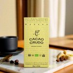 Obal čokolády Cacao Crudo s banánem, zobrazený na stole s kávovými zrny.